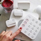 A Ring Alarm security kit is displayed on a small table with a hand pressing one of the buttons.