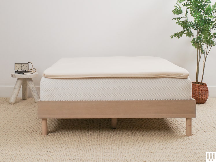 Birch Plush Organic Mattress Topper, a beige pad on top of a white mattress resting on a minimalist wooden bedframe with a small nightstand to the left and potted plant to the right
