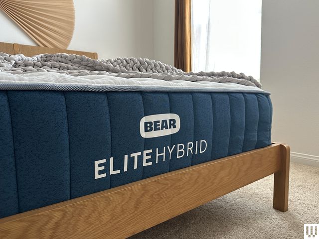 Closeup of the front end of a Bear Elite Hybrid Mattress with blue trim and white top, on a minimalist wooden bed frame