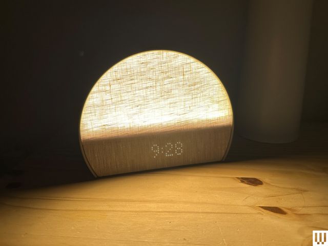 Hatch Restore 3, a semi-circular shaped alarm clock with an illuminated linen textured front, sitting on a wooden nightstand