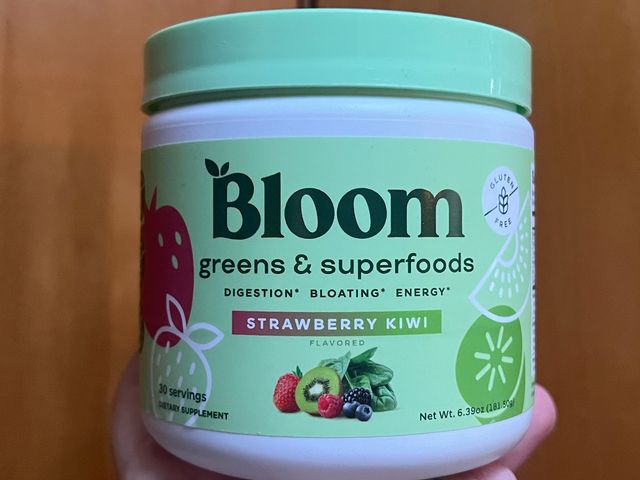 A hand holding up a small teal container of Bloom Nutrition Superfood Greens Powder