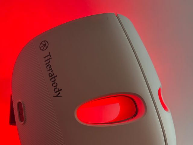 Therabody Theraface Mask, a white full facial mask with grey straps and inner panels illuminated with red lights