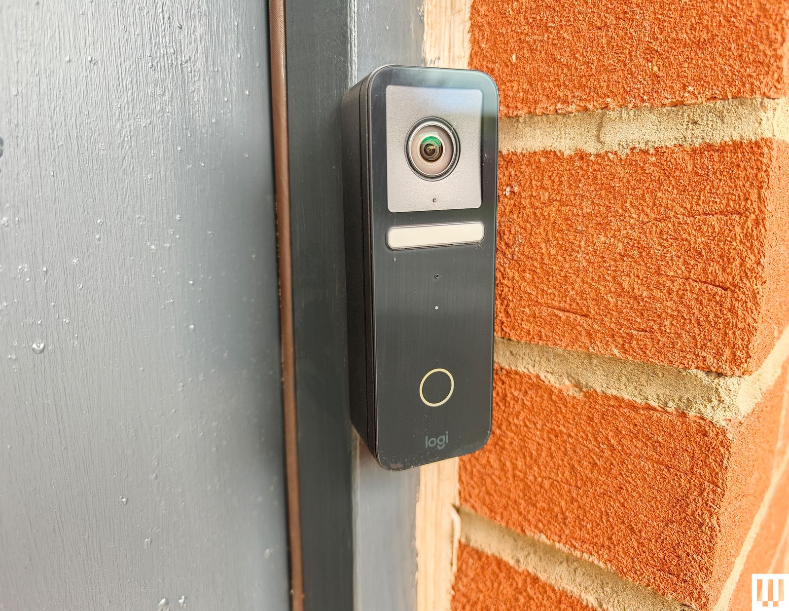 Black rectangular electronic doorbell with builtin camera attached to a grey door frame near a brick wall.