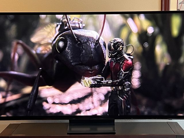Front view of Samsung S90D Q.D.-O.L.E.D., a large screen tv showing a movie scene of a miniature human in a special superhero suit standing beside a bug.