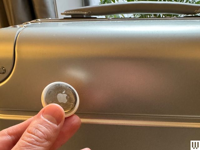Hand holding small silver circular disc with an engraved apple logo on it, in front of large silver luggage case