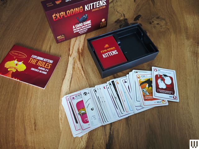Contents of a card game spread out on a wooden surface. In the back is the red box and pamphlet, in the center are the cards spread out in a line on top of each other, and to the right is the empty box.